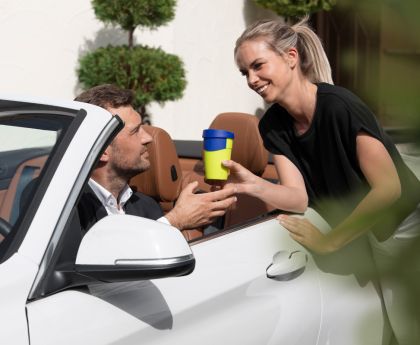 Preview: Eine Frau reicht ihrem Mann den Kaffeebecher ins Auto nach